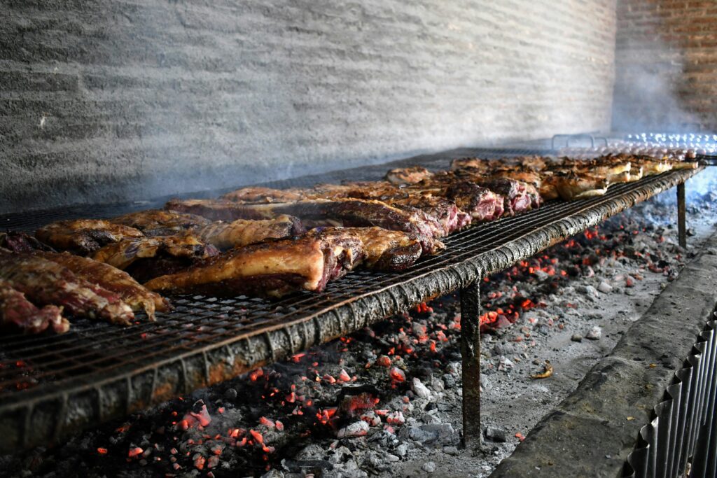 Grande quantidade de carne grelhando lentamente em uma parrilla (churrasqueira) tradicional, com brasas acesas, representando o autêntico churrasco argentino.